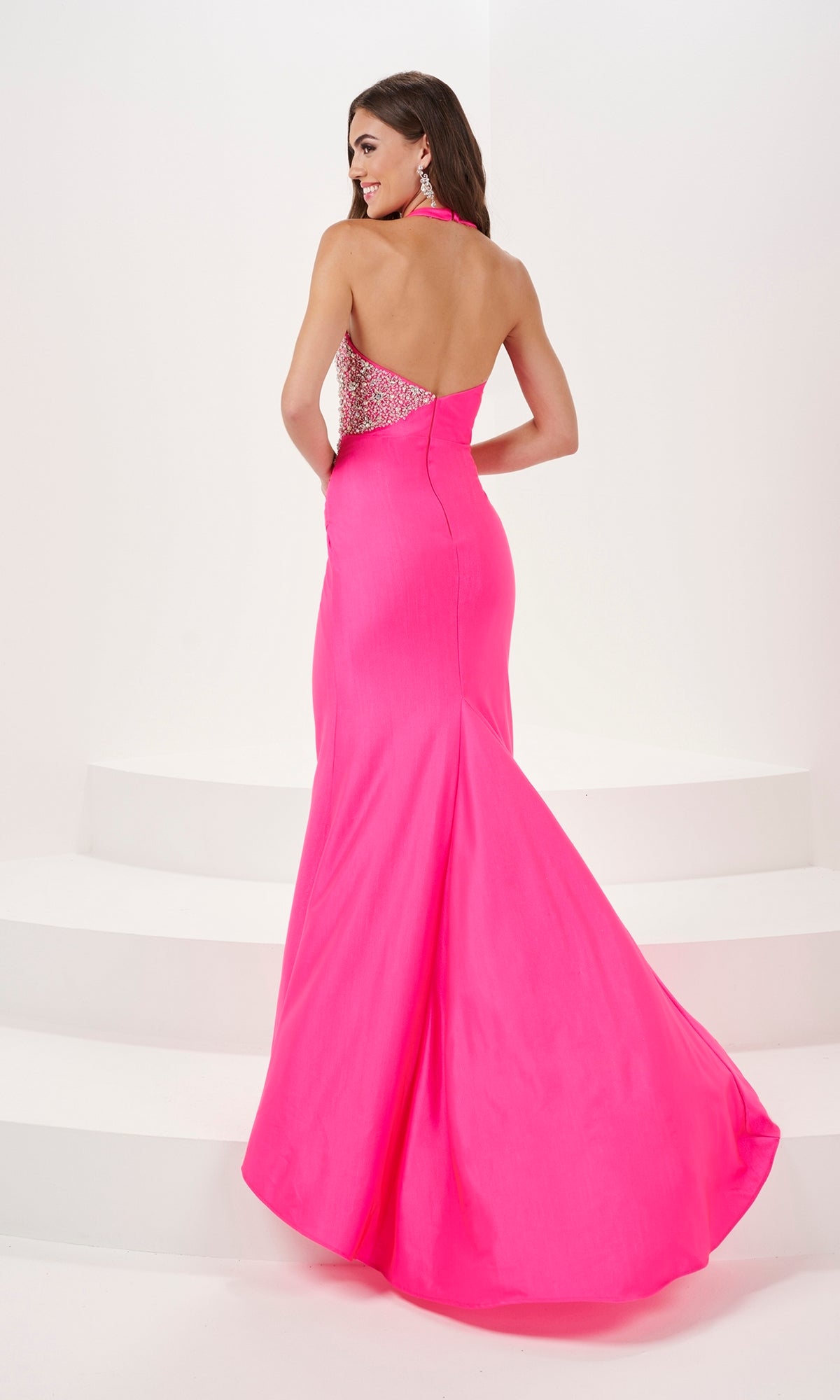 Woman in the Beaded-Bodice Draped Long Prom Dress 14175, featuring a bright pink hue, beaded sweetheart bodice, open back, and floor-length skirt, stands on white steps looking over her shoulder.